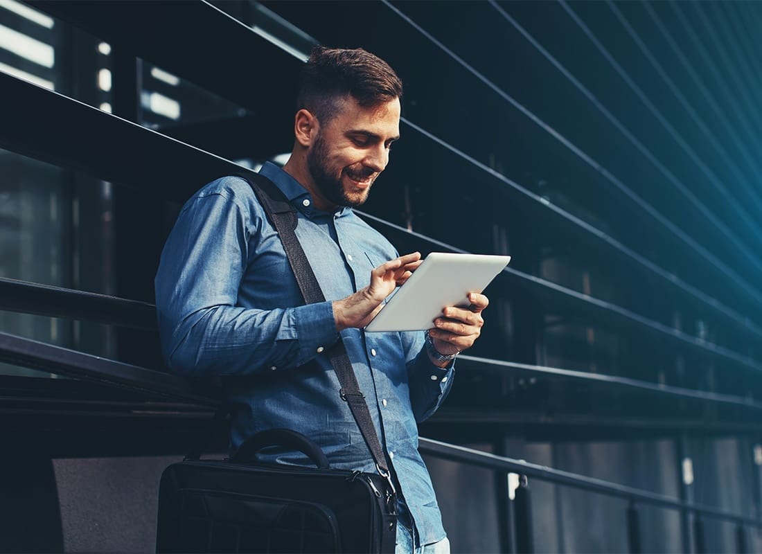 Blog - Young Smiling Business Man Standing Next to a Modern Building in the City While Holding and Using a White Tablet