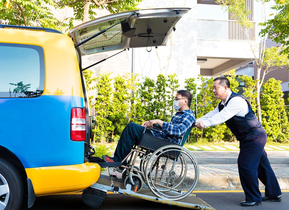 NEMT Insurance - Mature Driver Pushing a Young Man in a Wheelchair with a Mask into the Back of a Taxi Van