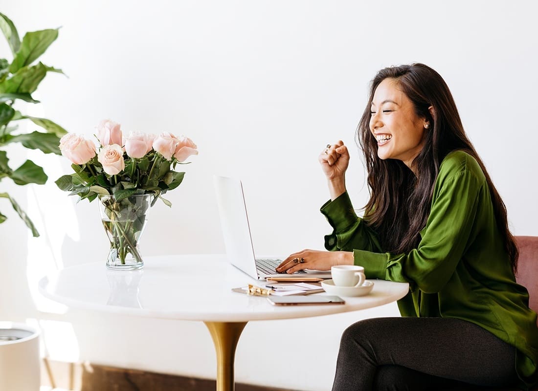 Service Center - Portrait of a Cheerful Young Business Woman Sitting at a Small Round Table with a Cup of Coffee and Pink Roses While Working on Her Laptop
